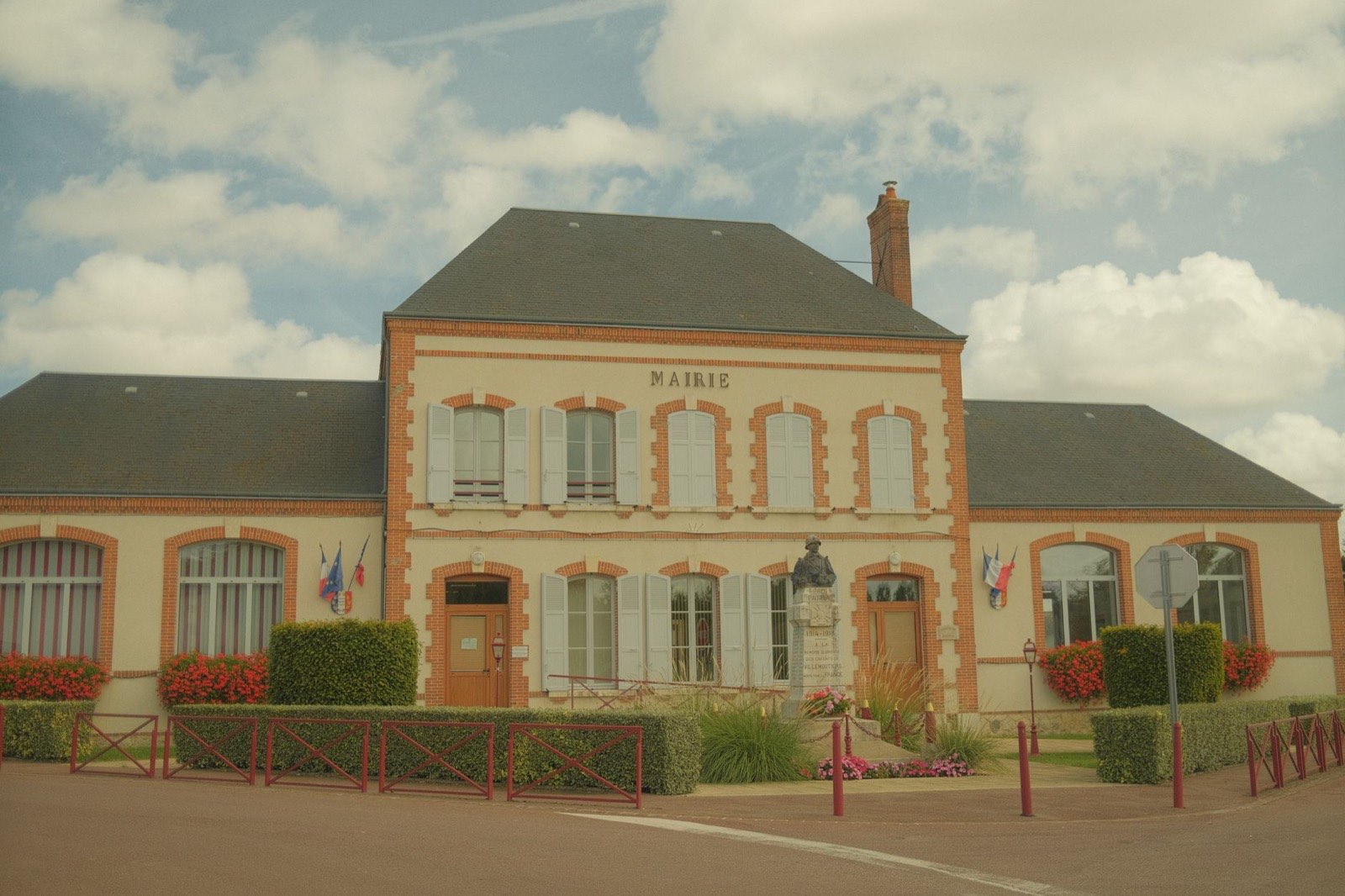 Façade d’une mairie de village, drapeaux tricolores encadrant l’entrée et monument aux morts sur le parvis.